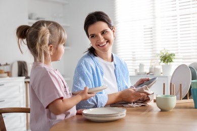 Photo of Little girl helping her mom wiping plates at table in kitchen