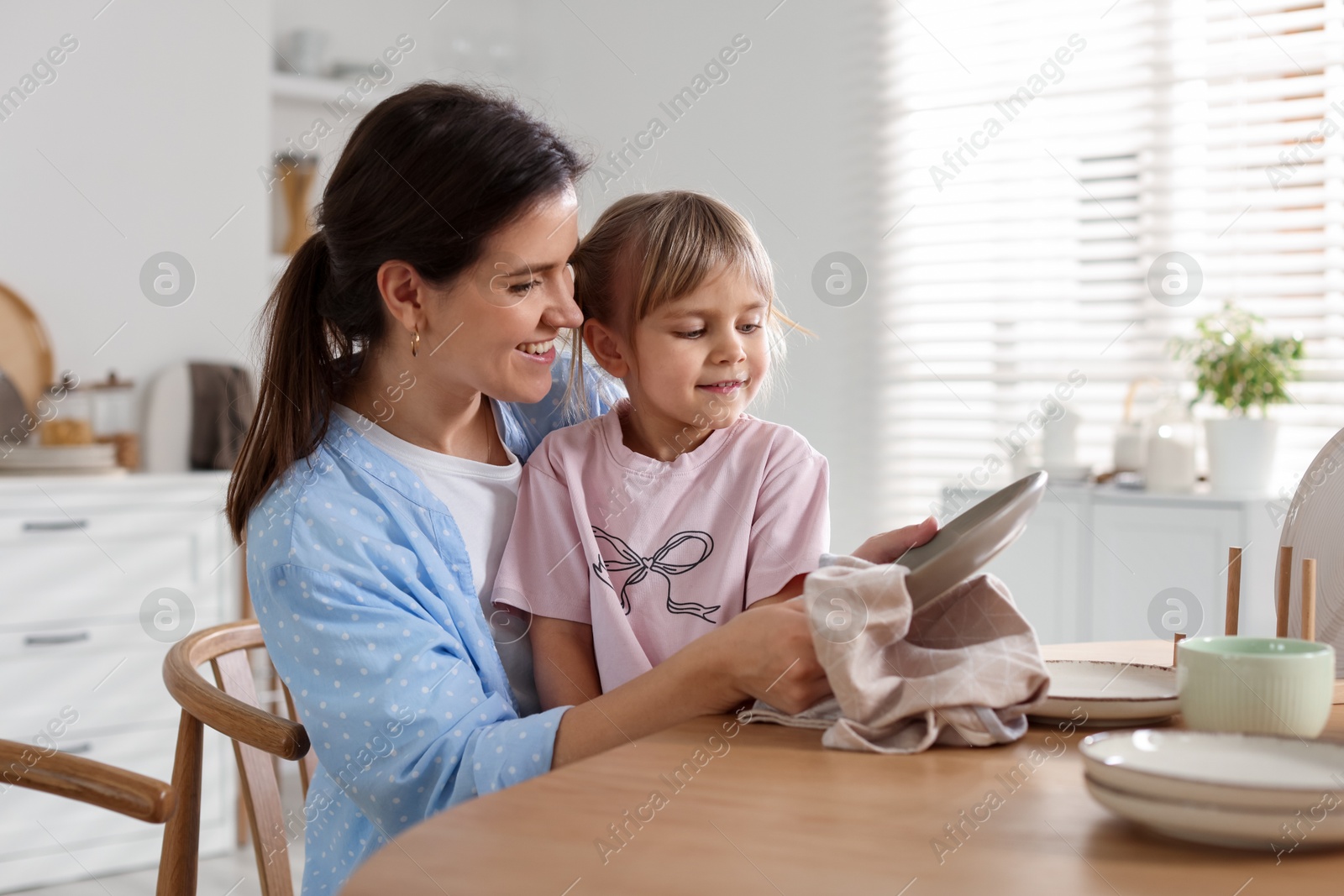 Photo of Little girl helping her mom wiping plates at table in kitchen