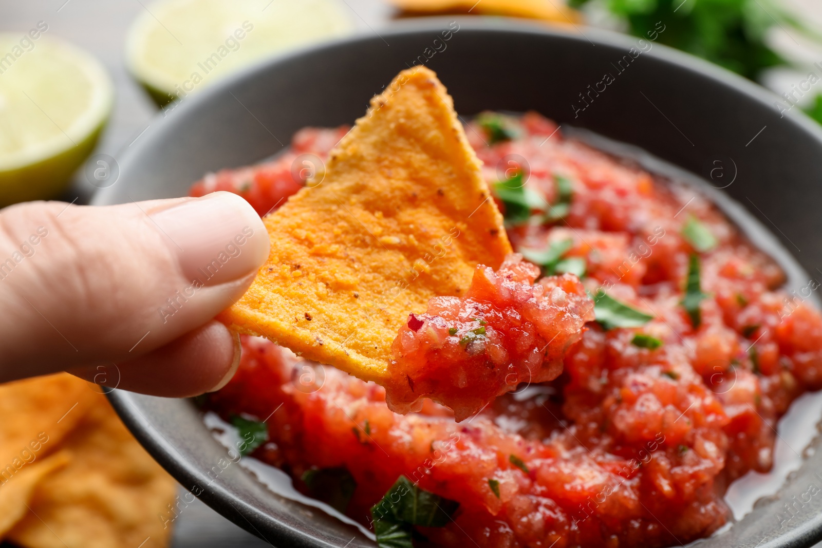 Photo of Woman dipping nacho chip into spicy salsa sauce at table, closeup