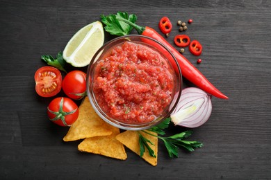 Spicy salsa sauce in bowl, nachos and ingredients on wooden table, flat lay