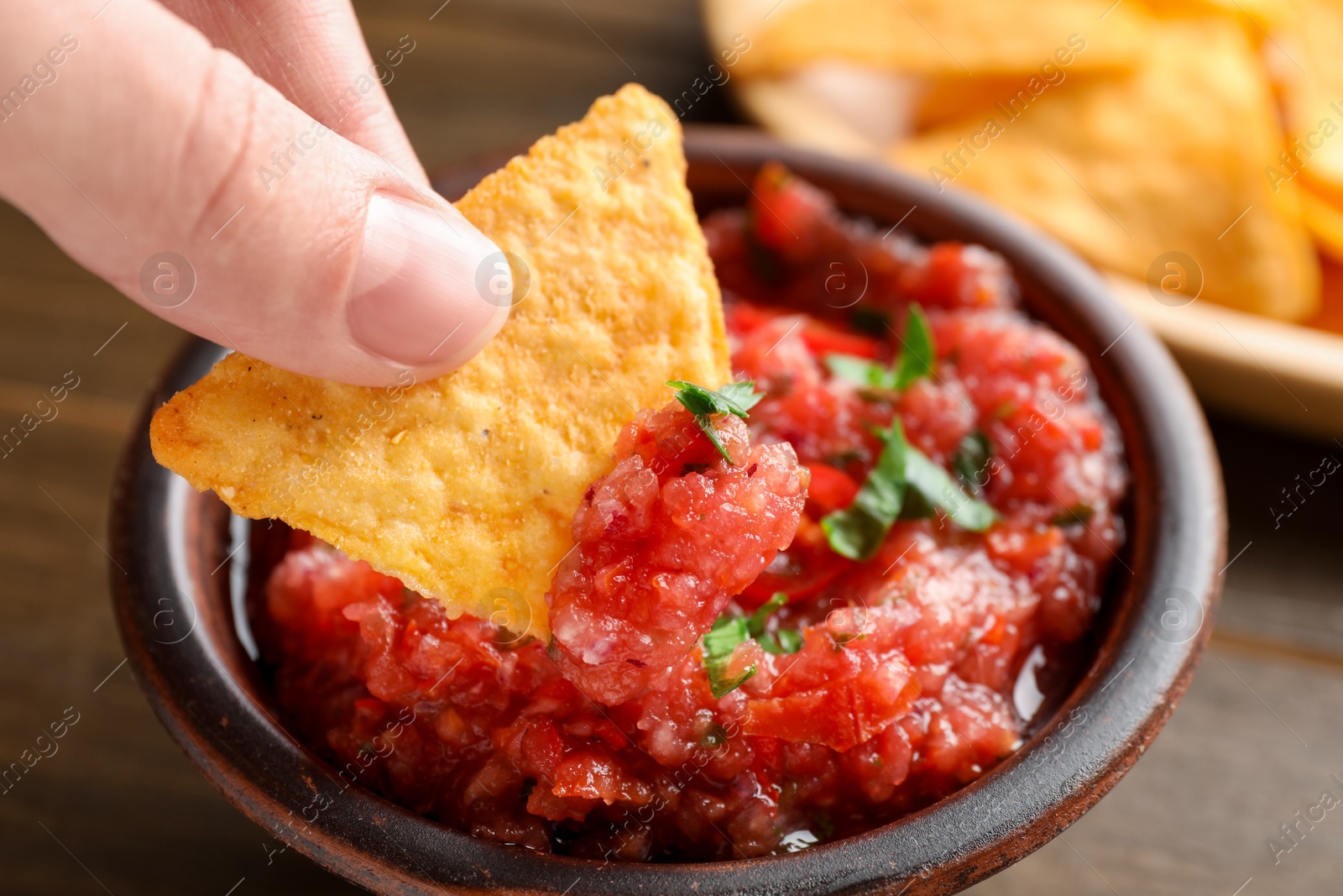 Photo of Woman dipping nacho chip into spicy salsa sauce at table, closeup