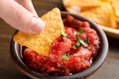 Photo of Woman dipping nacho chip into spicy salsa sauce at table, closeup