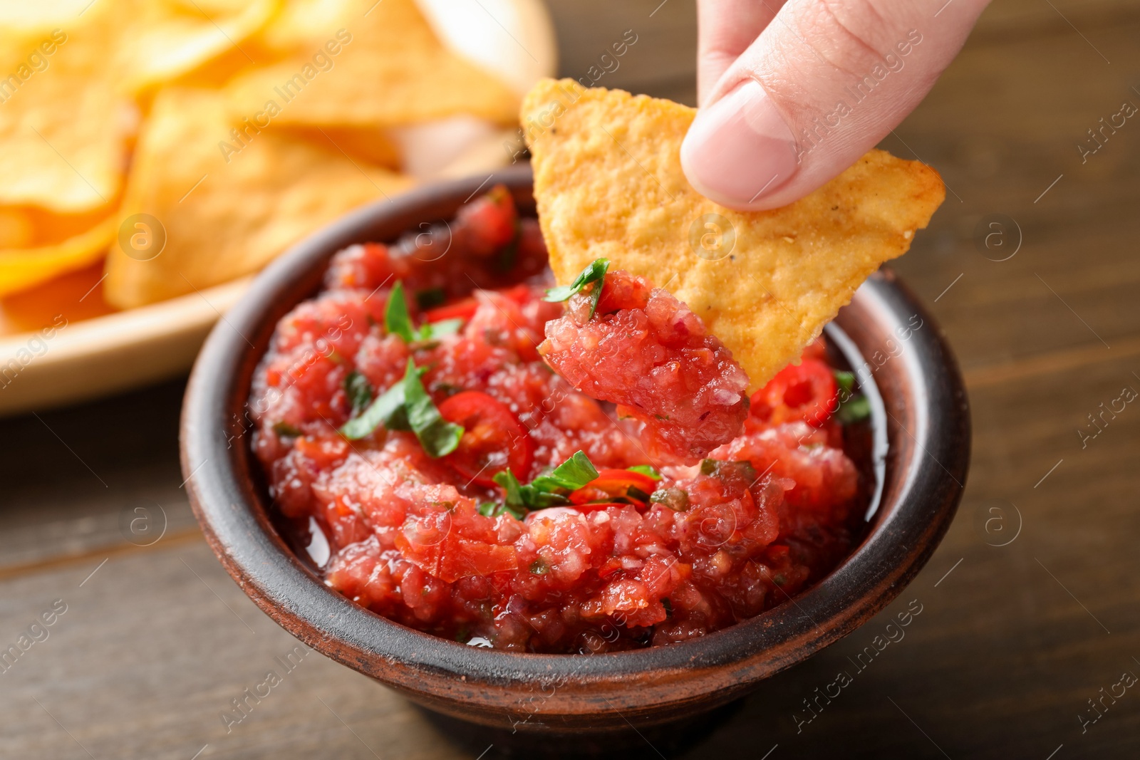 Photo of Woman dipping nacho chip into spicy salsa sauce at table, closeup