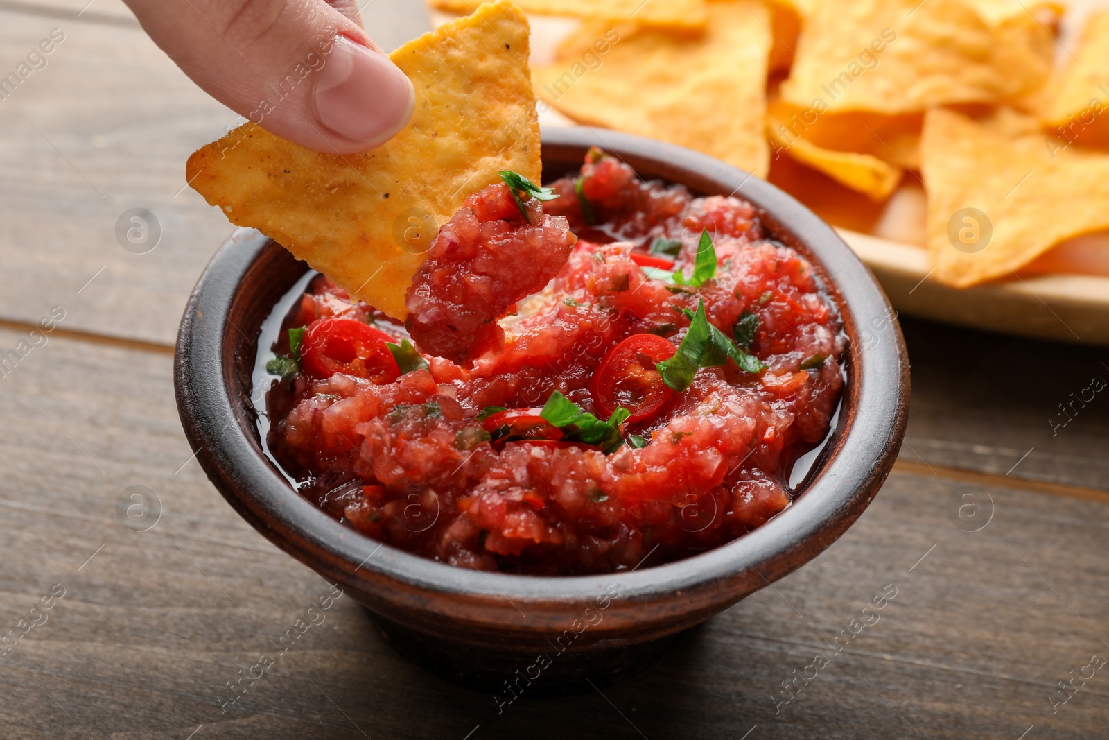 Photo of Woman dipping nacho chip into spicy salsa sauce at wooden table, closeup