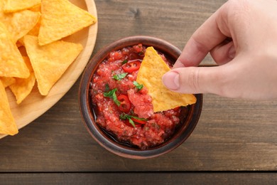 Photo of Woman dipping nacho chip into spicy salsa sauce at wooden table, top view