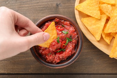 Woman dipping nacho chip into spicy salsa sauce at wooden table, top view