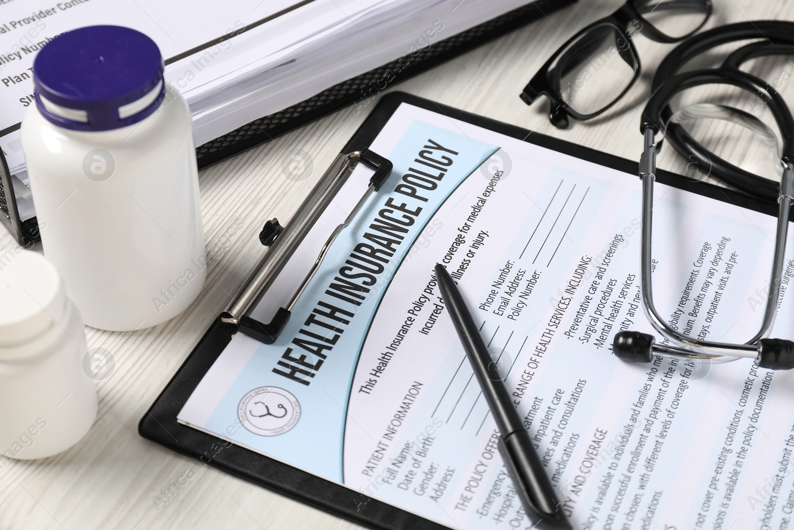 Photo of Health insurance policy form, stethoscope, glasses, pills and pen on light wooden table, closeup