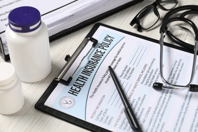 Health insurance policy form, stethoscope, glasses, pills and pen on light wooden table, closeup