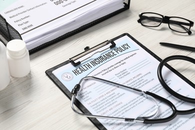 Health insurance policy form, medical cards, stethoscope, pills and pen on light wooden table, closeup