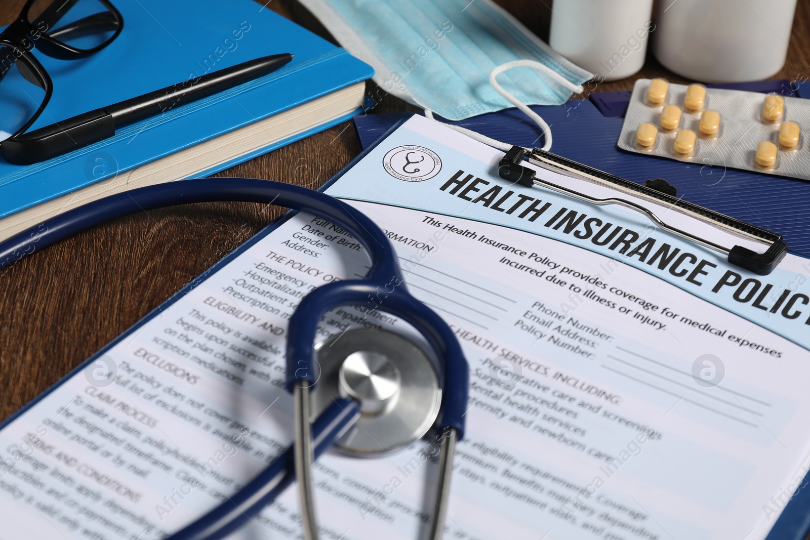 Photo of Health insurance policy form, stethoscope, notebook, pills and pen on wooden table, closeup