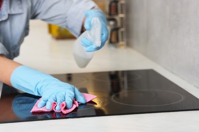 Photo of Woman using cleaning product while wiping induction cooktop with rag in kitchen, closeup