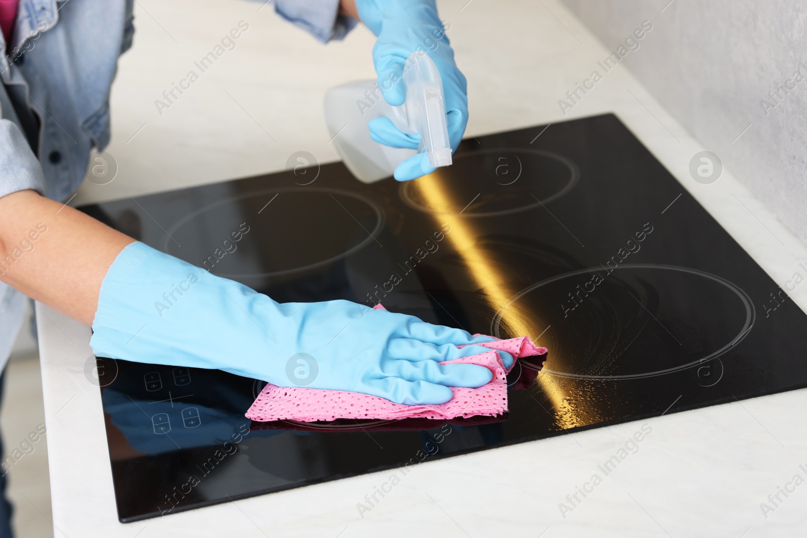 Photo of Woman using cleaning product while wiping induction cooktop with rag in kitchen, closeup