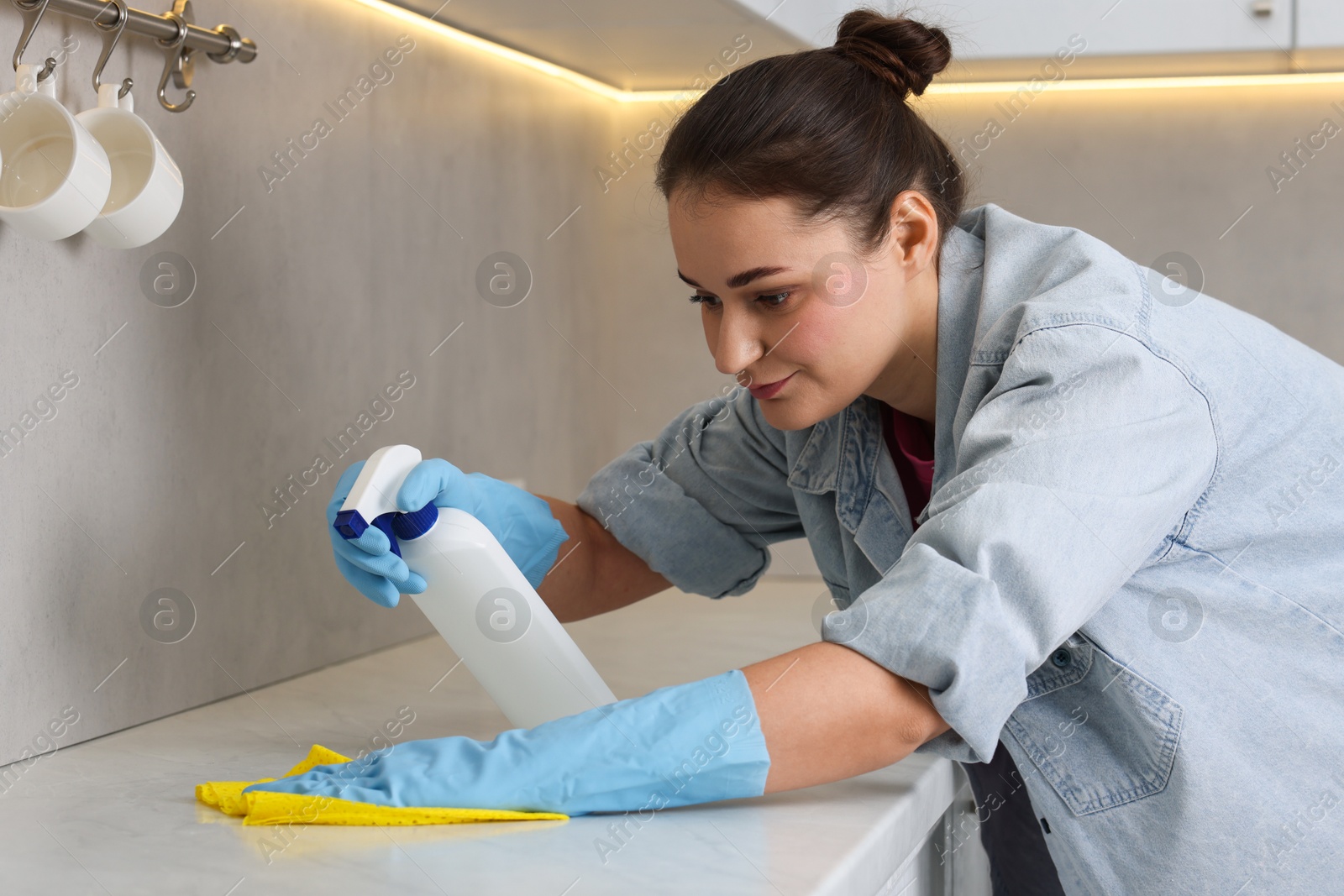 Photo of Woman using cleaning product while wiping countertop with rag indoors