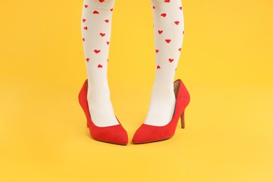 Photo of Little girl wearing oversized high heeled shoes on orange background, closeup