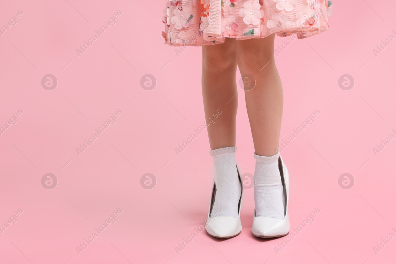 Photo of Little girl wearing oversized high heeled shoes on pink background, closeup. Space for text