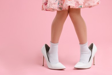 Photo of Little girl wearing oversized high heeled shoes on pink background, closeup. Space for text