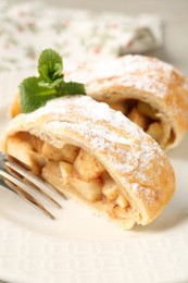 Pieces of tasty apple strudel with powdered sugar, mint and fork on plate, closeup