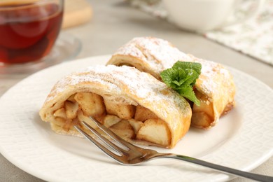 Pieces of tasty apple strudel with powdered sugar, mint and fork on grey textured table, closeup