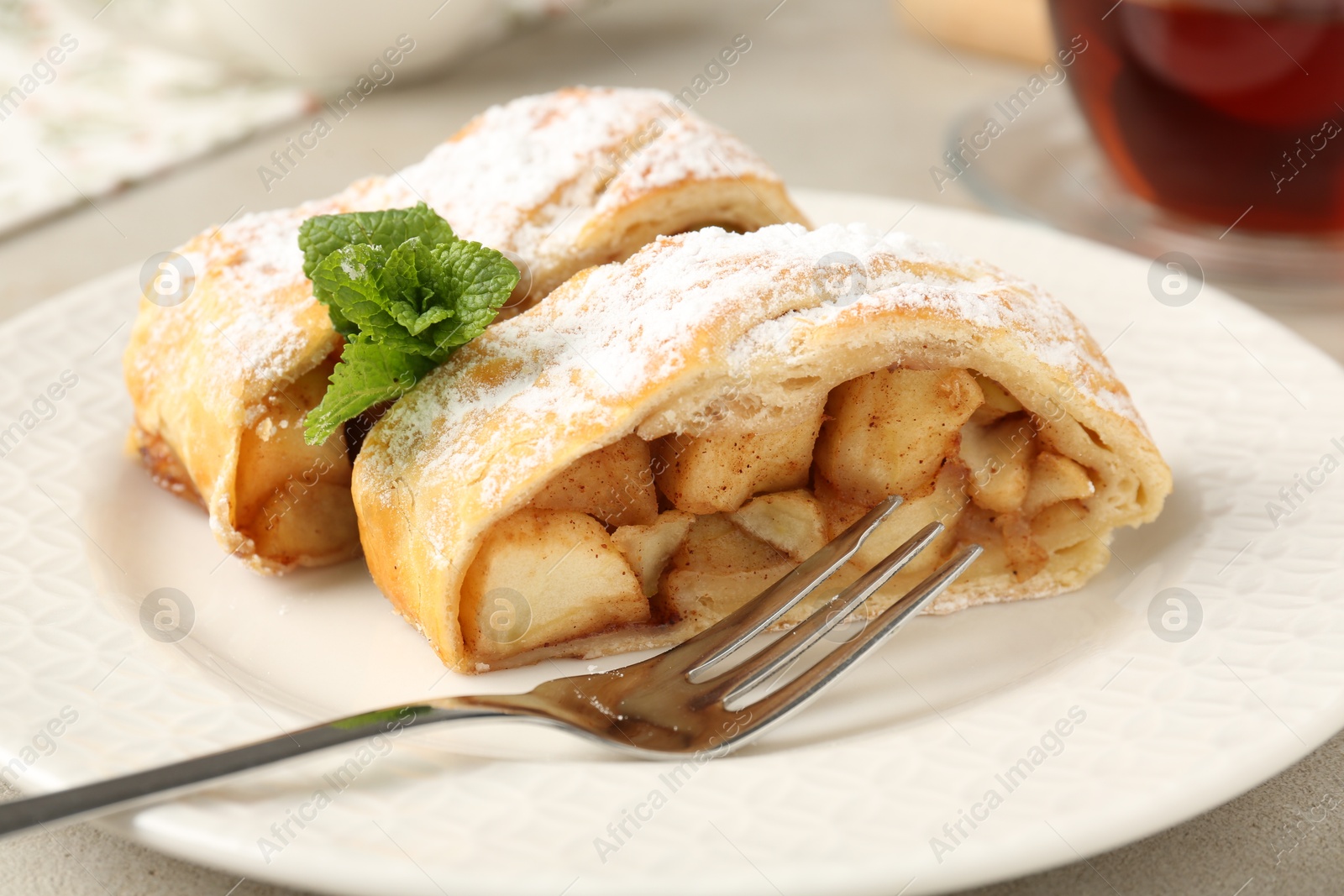 Photo of Pieces of tasty apple strudel with powdered sugar, mint and fork on grey textured table, closeup