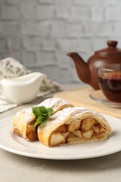 Photo of Pieces of tasty apple strudel with powdered sugar, mint and fork on grey textured table, closeup