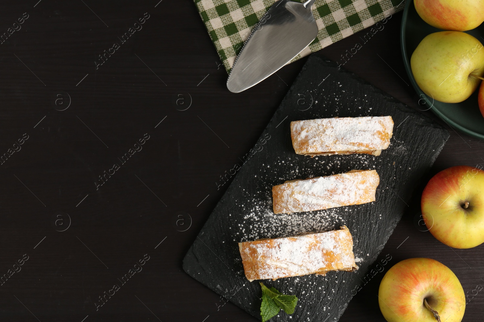 Photo of Pieces of tasty apple strudel with powdered sugar, mint and fruits on black wooden table, flat lay. Space for text