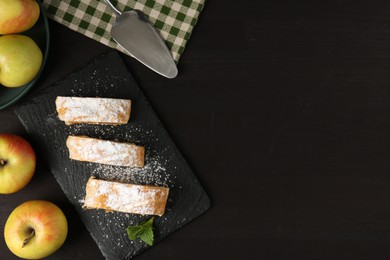 Photo of Pieces of tasty apple strudel with powdered sugar, mint and fruits on black wooden table, flat lay. Space for text