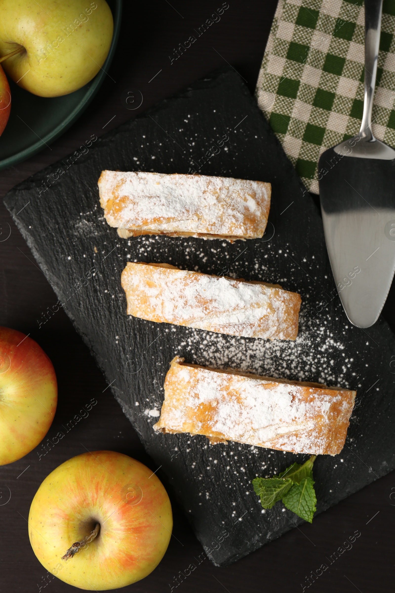 Photo of Pieces of tasty apple strudel with powdered sugar, mint and fruits on black wooden table, flat lay