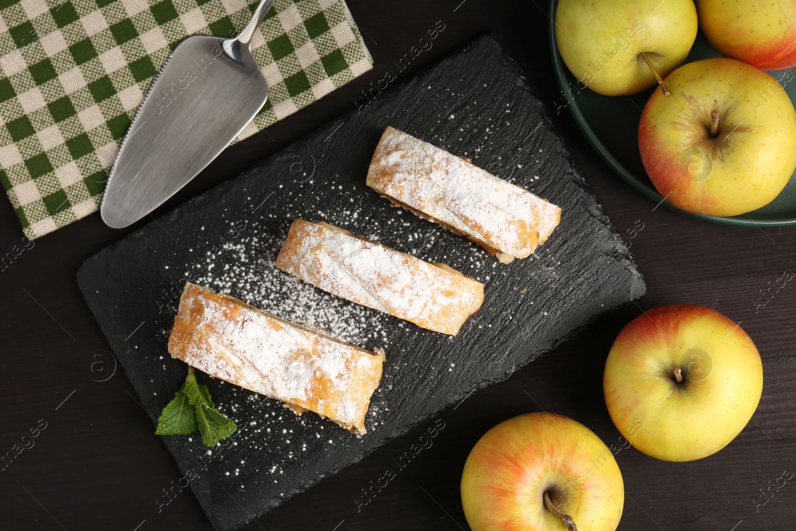 Photo of Pieces of tasty apple strudel with powdered sugar, mint and fruits on black wooden table, flat lay