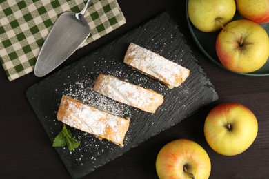 Photo of Pieces of tasty apple strudel with powdered sugar, mint and fruits on black wooden table, flat lay