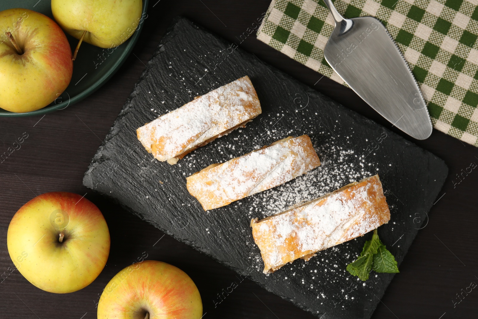 Photo of Pieces of tasty apple strudel with powdered sugar, mint and fruits on black wooden table, flat lay