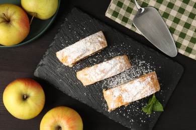 Photo of Pieces of tasty apple strudel with powdered sugar, mint and fruits on black wooden table, flat lay