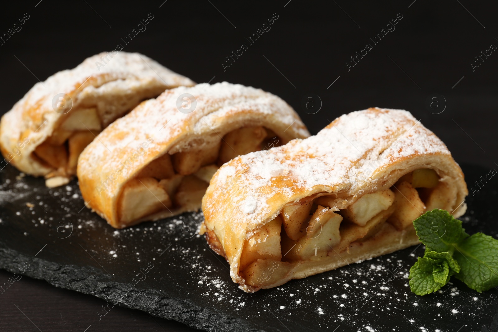 Photo of Pieces of tasty apple strudel with powdered sugar and mint on black wooden table, closeup