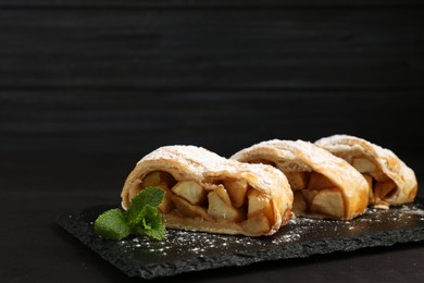 Photo of Pieces of tasty apple strudel with powdered sugar and mint on black table, closeup. Space for text