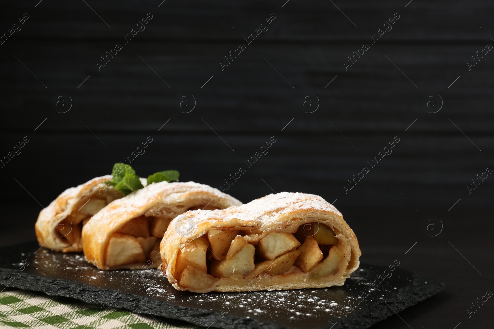 Photo of Pieces of tasty apple strudel with powdered sugar and mint on black table, closeup. Space for text