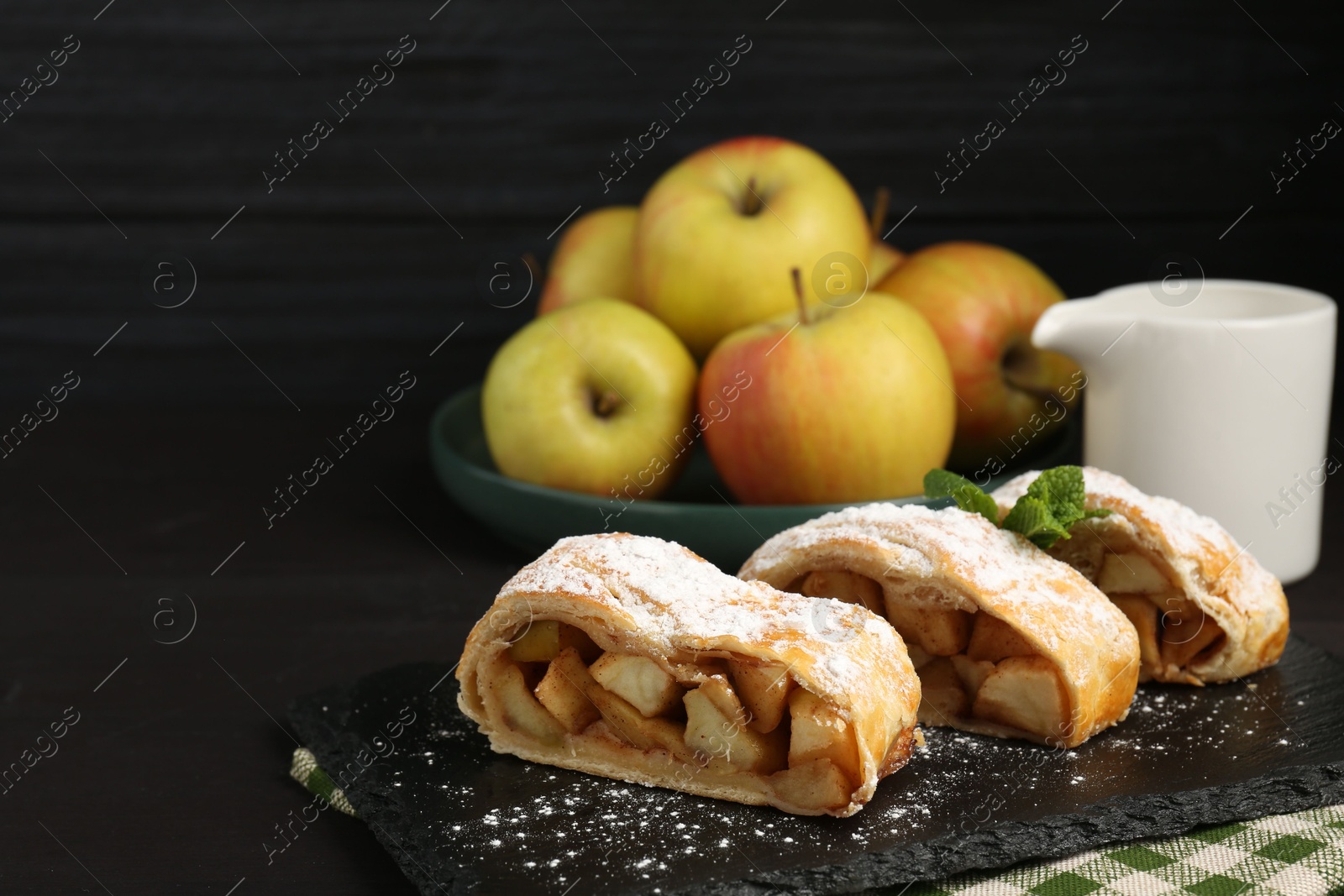 Photo of Pieces of tasty apple strudel with powdered sugar and mint on black table, closeup. Space for text