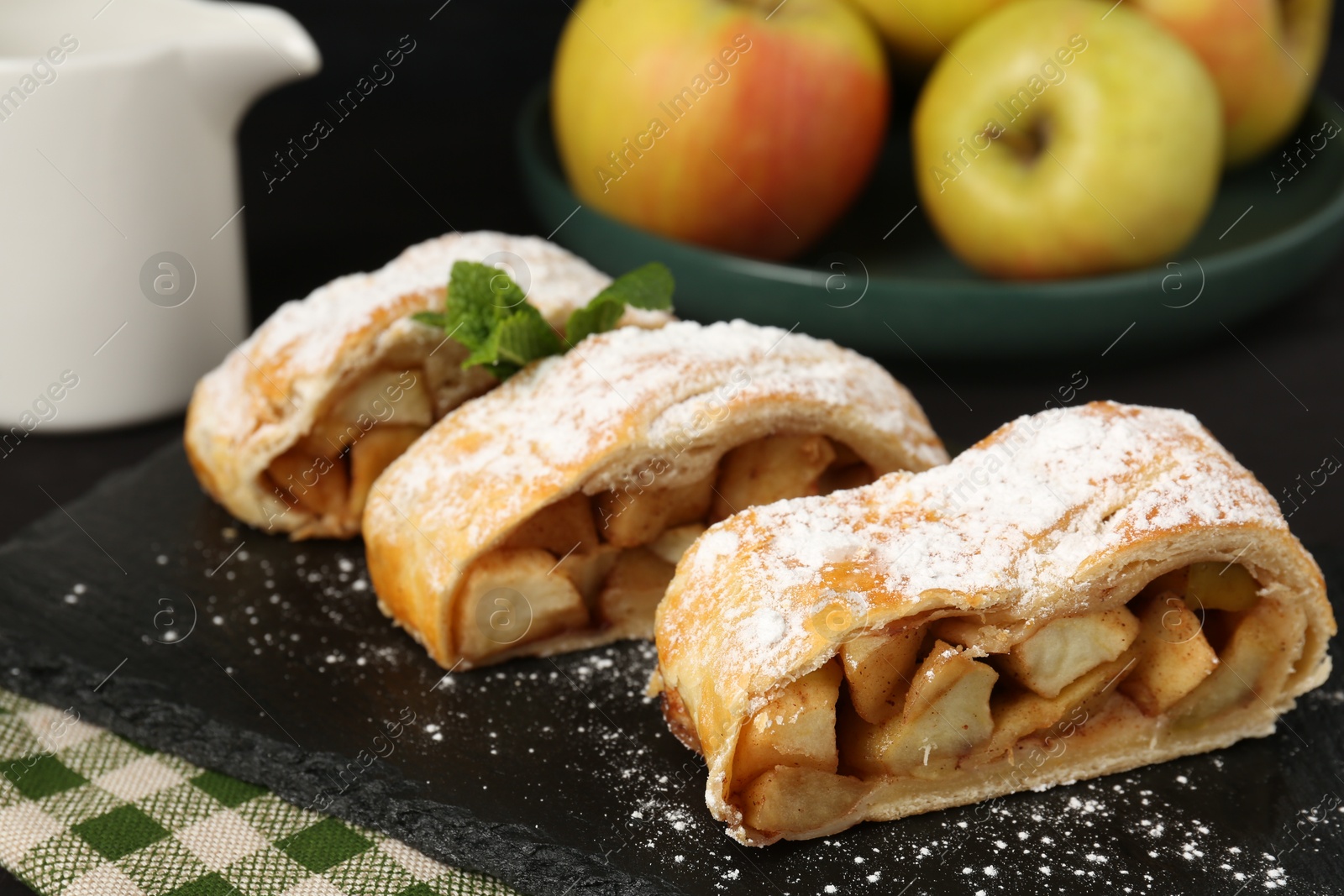 Photo of Pieces of tasty apple strudel with powdered sugar and mint on black table, closeup