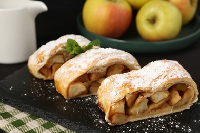 Pieces of tasty apple strudel with powdered sugar and mint on black table, closeup