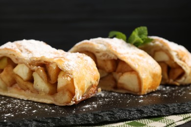 Photo of Pieces of tasty apple strudel with powdered sugar and mint on table against black background, closeup