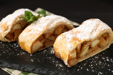 Photo of Pieces of tasty apple strudel with powdered sugar and mint on table against black background, closeup