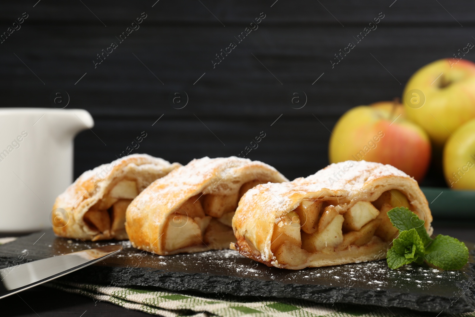 Photo of Pieces of tasty apple strudel with powdered sugar and mint on table against black background, closeup