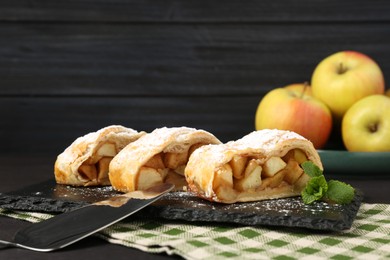 Photo of Pieces of tasty apple strudel with powdered sugar and mint on black table, closeup