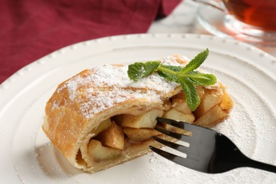 Photo of Piece of tasty apple strudel with powdered sugar, mint and fork on table, closeup