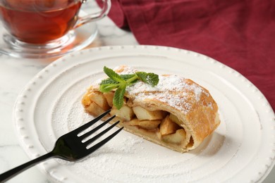 Photo of Piece of tasty apple strudel with powdered sugar, mint and fork on white table, closeup