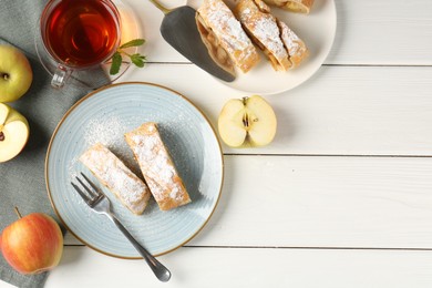 Photo of Pieces of tasty apple strudel with powdered sugar, fruits and tea on white wooden table, flat lay. Space for text