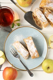 Photo of Pieces of tasty apple strudel with powdered sugar, fruits and tea on white wooden table, flat lay