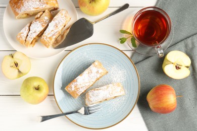 Photo of Pieces of tasty apple strudel with powdered sugar, fruits and tea on white wooden table, flat lay