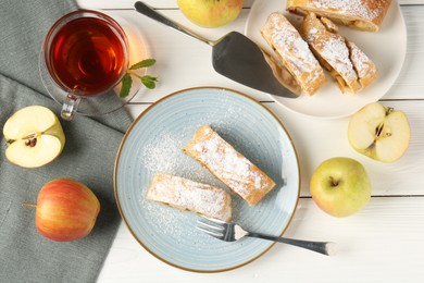 Photo of Pieces of tasty apple strudel with powdered sugar, fruits and tea on white wooden table, flat lay