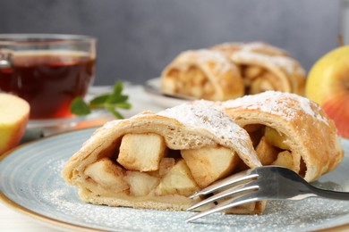 Photo of Pieces of tasty apple strudel with powdered sugar and fork on white table against grey background, closeup