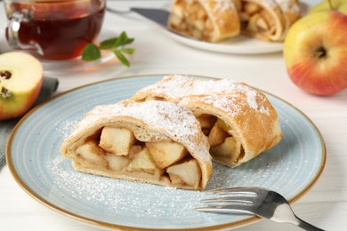 Photo of Pieces of tasty apple strudel with powdered sugar and fork on white wooden table, closeup