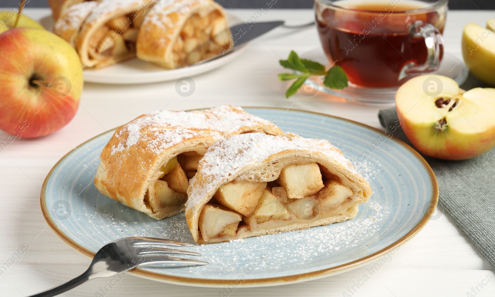 Photo of Pieces of tasty apple strudel with powdered sugar and fork on white wooden table against grey background, closeup